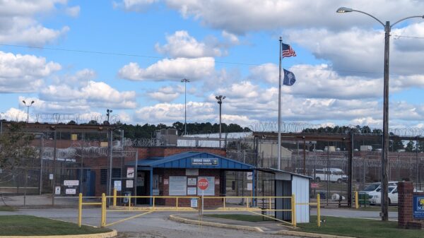 Entrance of the Broad River Correctional Institution with razor wire, fences, and the South Carolina and US flags under a blue sky with white clouds