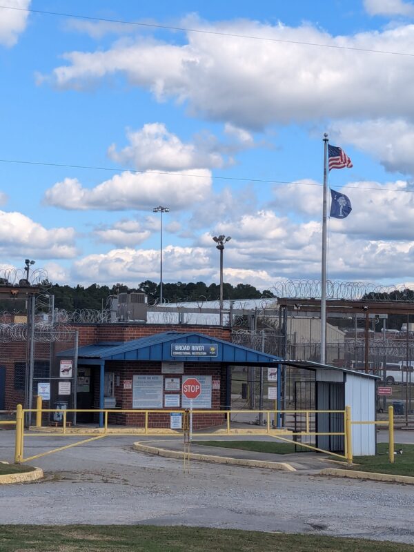 Entrance of the Broad River Correctional Institution with razor wire, fences, and the South Carolina and US flags under a blue sky with white clouds