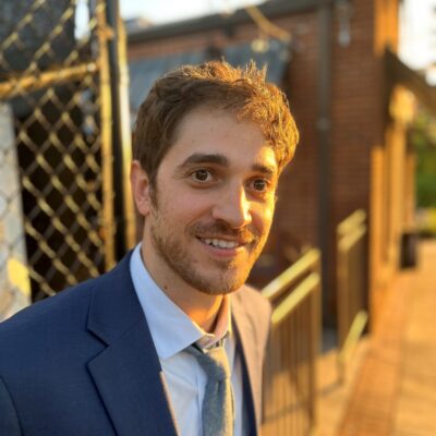 A photo of Josh Malkin in a  navy blue suit standing in the sunlight beside a chain link fence