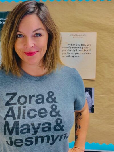 A photo of Mary Wood. She has shoulder-length brown hair and is wearing a gray T-shirt with the names "Zora & Alice & Maya & Jesmyn" on it.