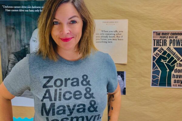 A photo of Mary Wood. She has shoulder-length brown hair and is wearing a gray T-shirt with the names "Zora & Alice & Maya & Jesmyn" on it.