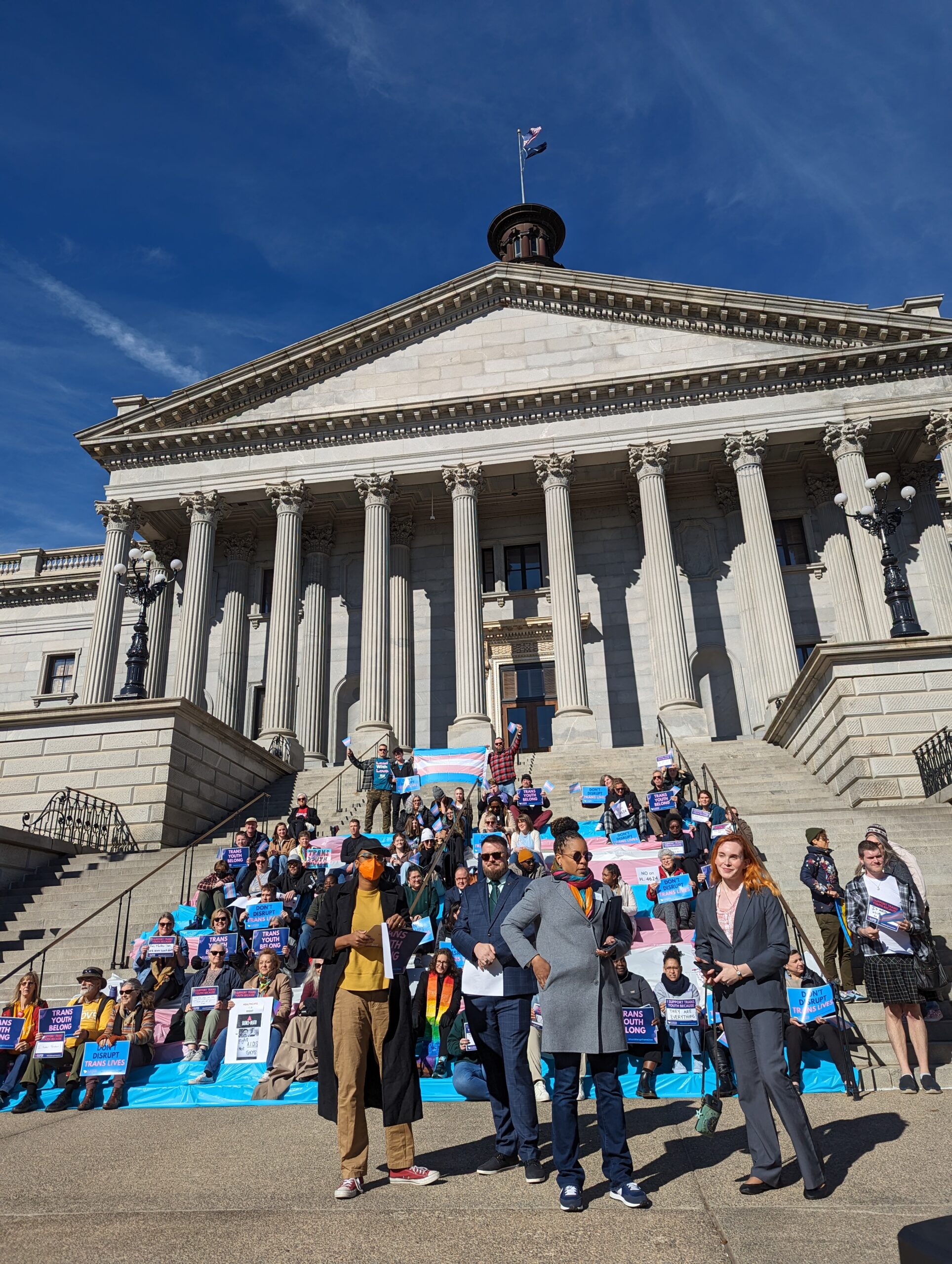 A crowd waving trans pride flags assembles on the State House steps