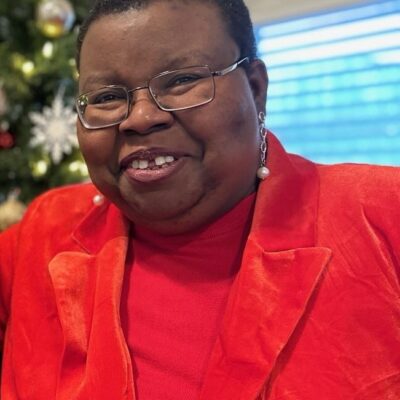 Tish Gotell Faulks in a bright red suit jacket and red shirt. She has a close cropped haircut, wire-rimmed glasses, and pearl earrings.