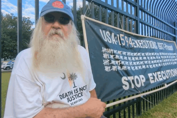 A man with a long white beard sits beside a black banner that reads, "USA: 1594 Executions 1977 - Present. Stop Executions!"