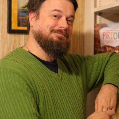 Photo of Matt standing beside a book case. He is smiling slightly and wearing a green cable knit sweater.