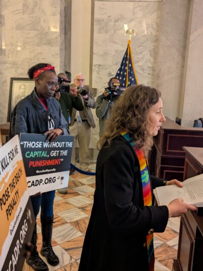 Petitioners, including a white woman in minister's garments and a Black man in a dress shirt, deliver petitions at a desk in the State House lobby.