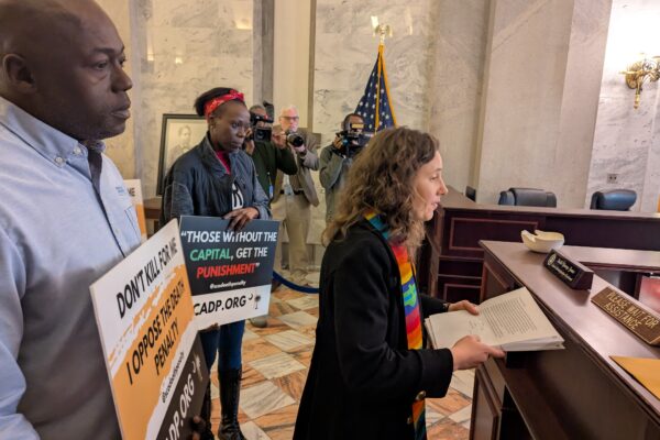 Petitioners, including a white woman in minister's garments and a Black man in a dress shirt, deliver petitions at a desk in the State House lobby.