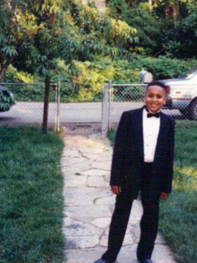 A young Black boy stands in a front yard smiling and wearing a tuxedo