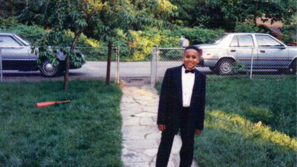 A young Black boy stands in a front yard smiling and wearing a tuxedo