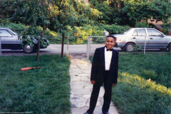 A young Black boy stands in a front yard smiling and wearing a tuxedo