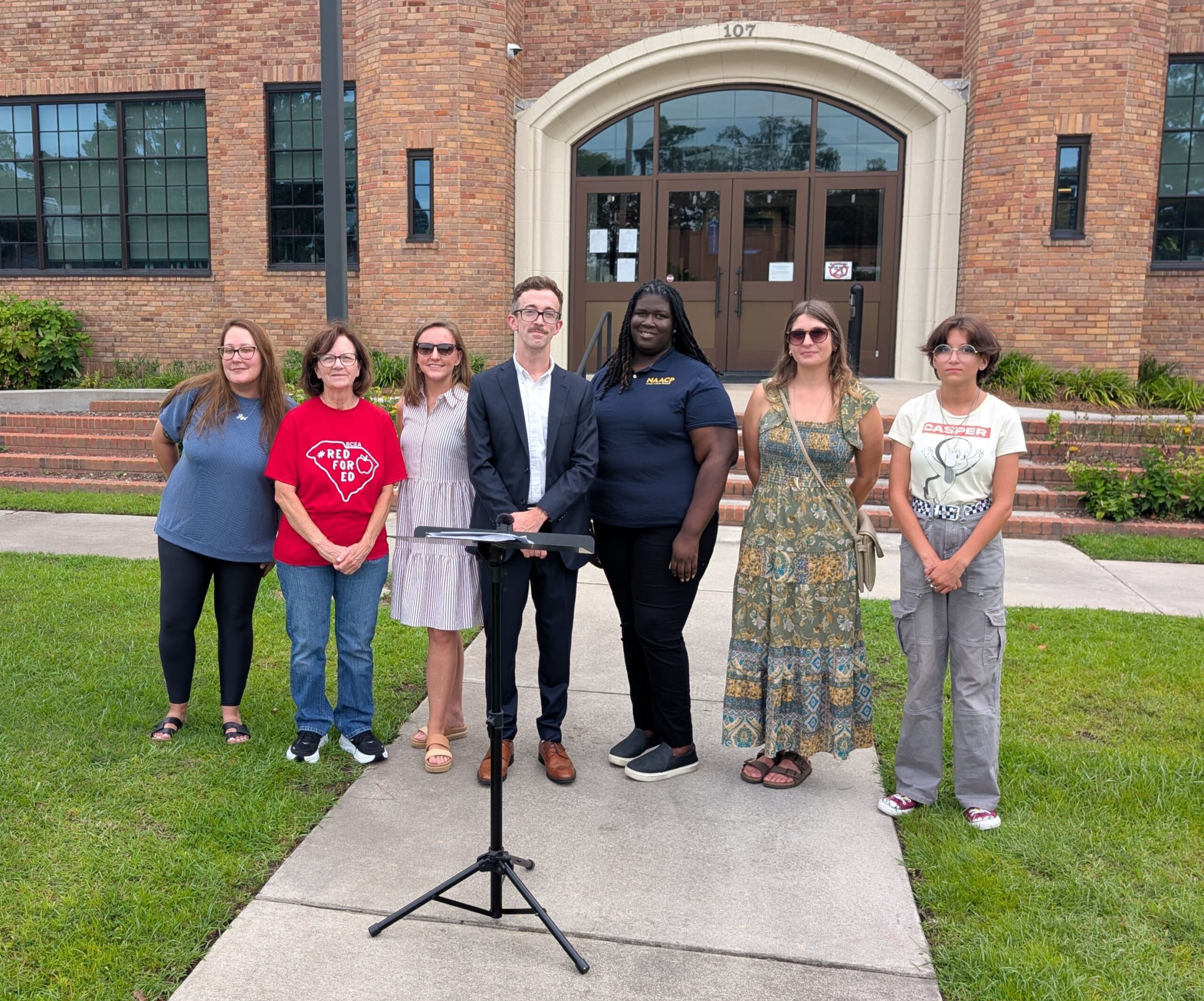 Seven speakers stand behind a metal lectern in front of the Berkeley County School District office, a large brick building.