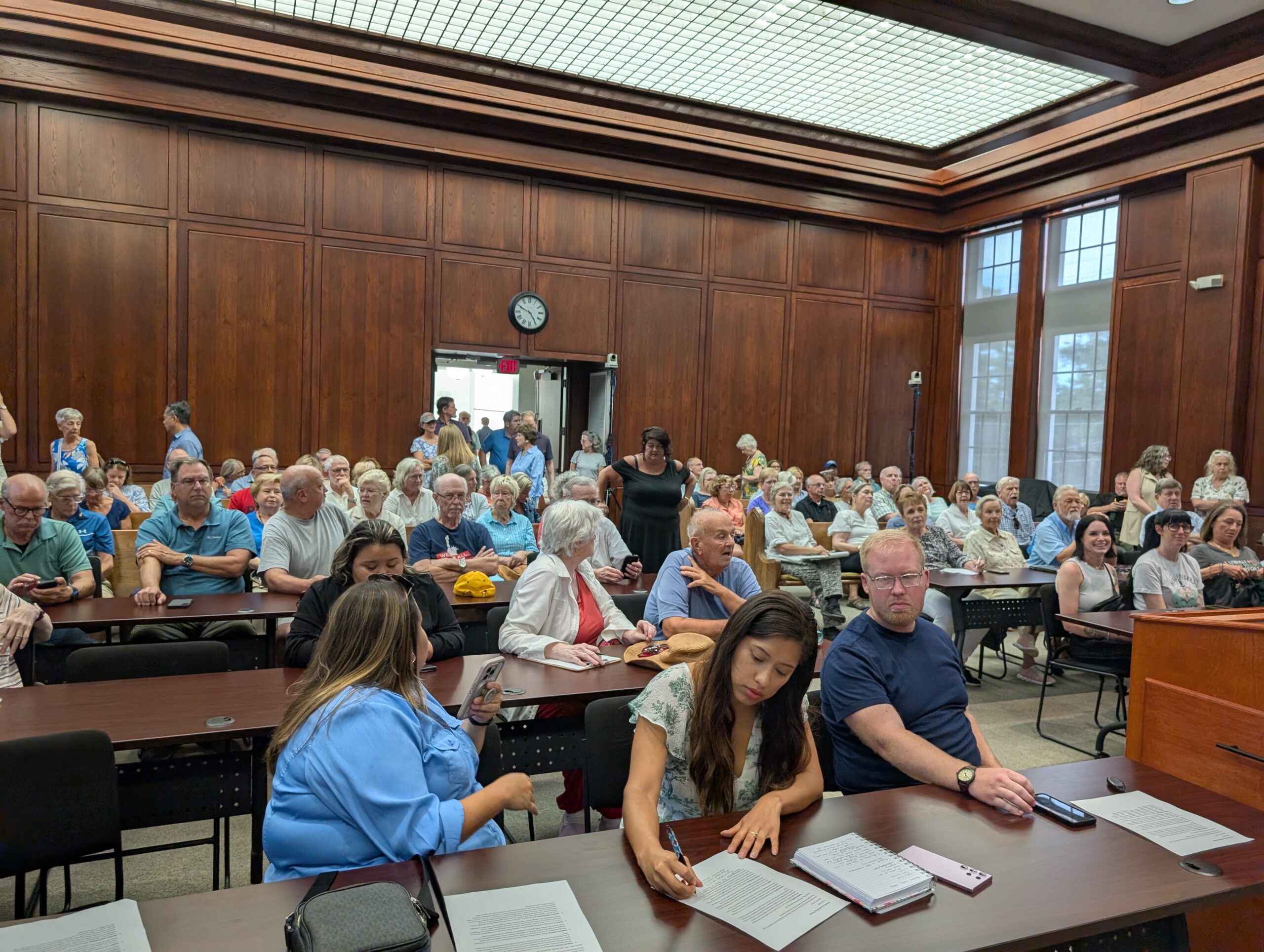 A crowd of people in a wood paneled room, seated in chairs and standing at the back