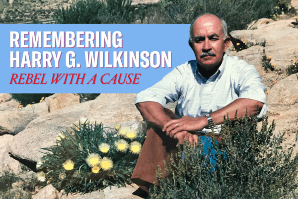 "Remembering Harry G. Wilkinson, Rebel with a Cause." Photo of Harry, a man in a button-up shirt with the sleeves rolled up, seated on some rocks with an outcropping of flowering scrub plants.