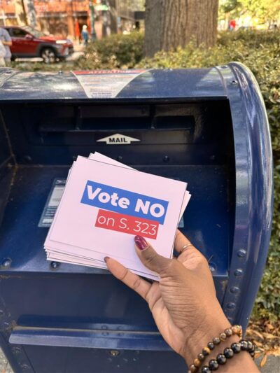 A hand puts a stack of postcards with the words "Vote No on S. 323" into a mailbox.