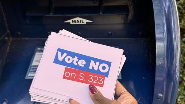 A hand puts a stack of postcards with the words "Vote No on S. 323" into a mailbox.