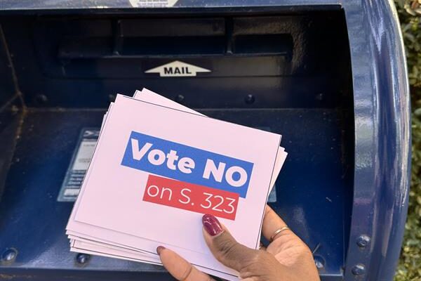 A hand puts a stack of postcards with the words "Vote No on S. 323" into a mailbox.