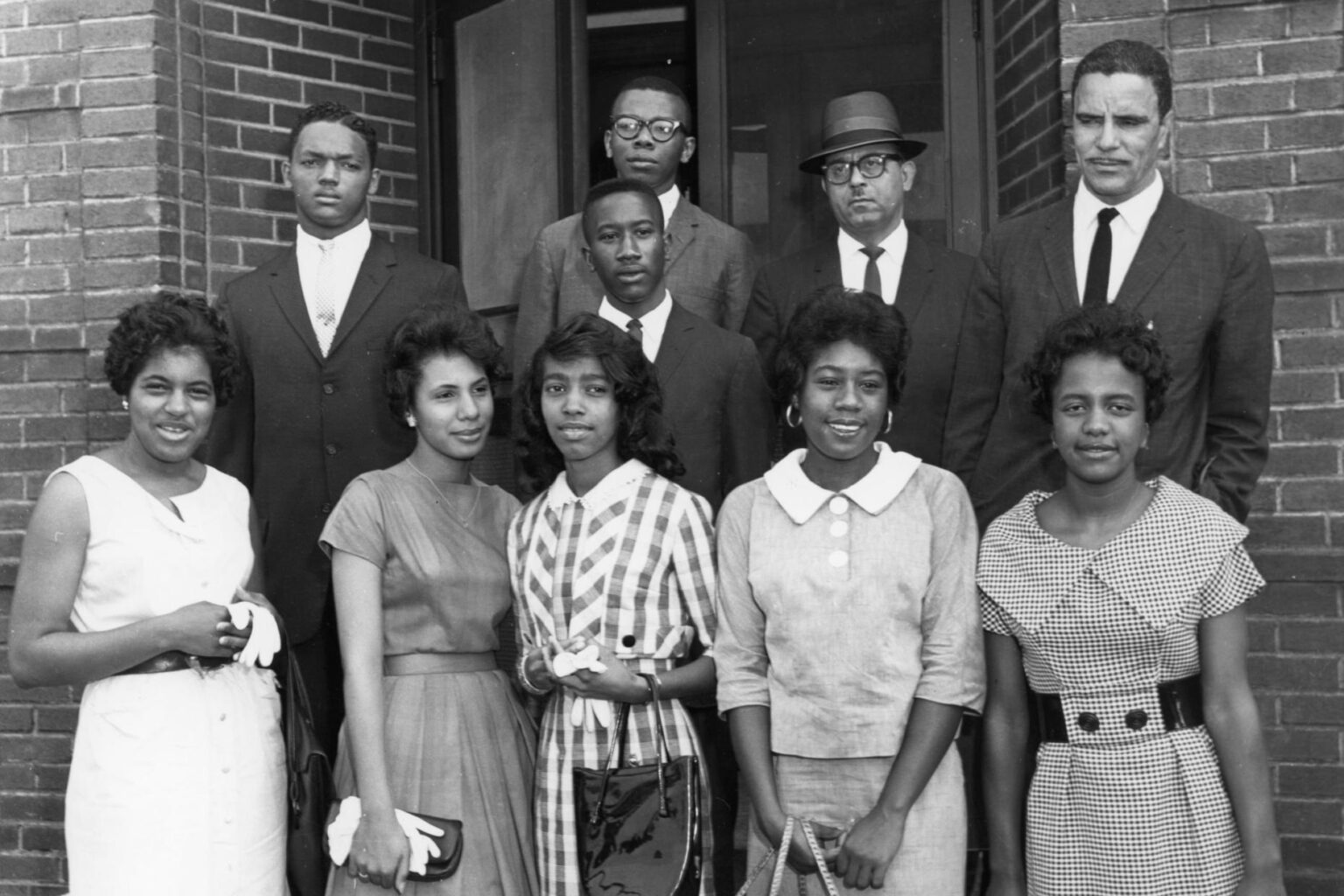 Black and white photo of eight students and two adults in suits standing in front of a brick building