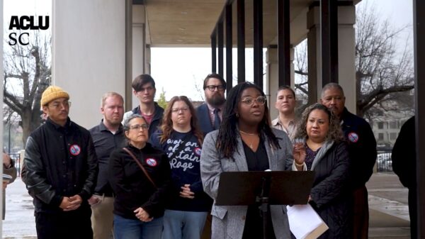 Photo of a press conference outside a brutalist Statehouse office building. Participants are wearing stickers that have the number 4764 crossed out.