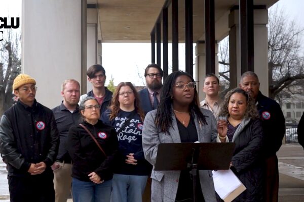 Photo of a press conference outside a brutalist Statehouse office building. Participants are wearing stickers that have the number 4764 crossed out.