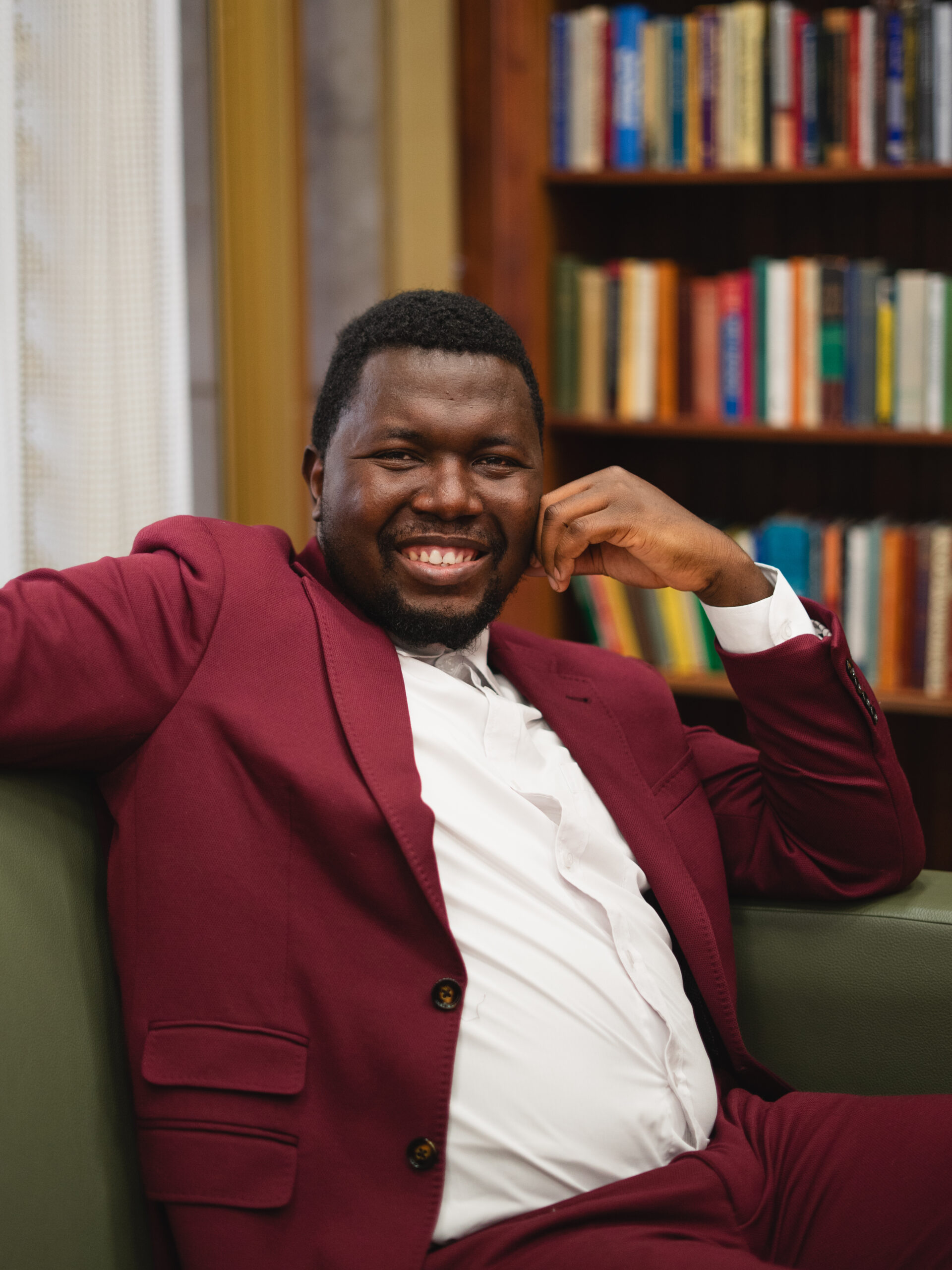 Matthew Ariwoola smiles while seated in a library. He is wearing a garnet suit jacket and white dress shirt.