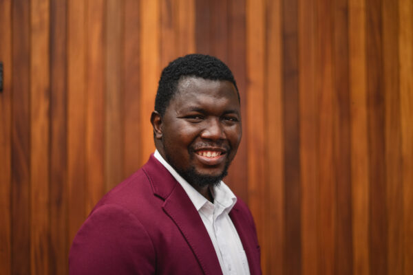 Matthew Ariwoola smiling while wearing a garnet suit jacket in front of a wood paneled wall