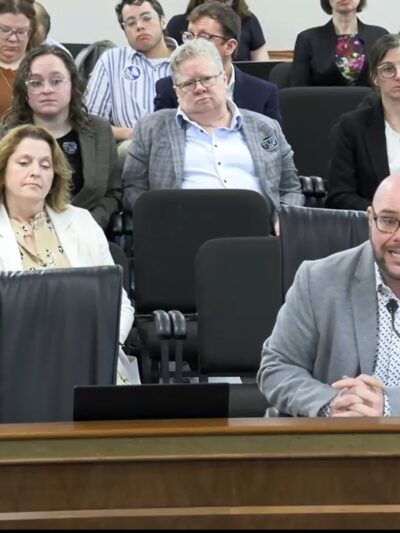 Jace Woodrum, a white man in a gray suit, speaks into a microphone in a Statehouse committee room with a crowd of people sitting behind him