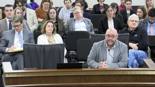 Jace Woodrum, a white man in a gray suit, speaks into a microphone in a Statehouse committee room with a crowd of people sitting behind him
