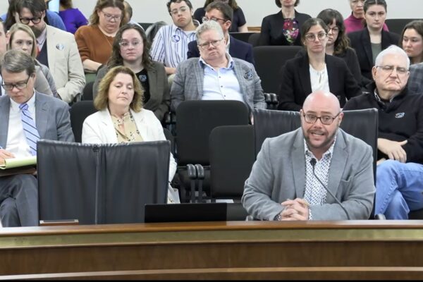 Jace Woodrum, a white man in a gray suit, speaks into a microphone in a Statehouse committee room with a crowd of people sitting behind him