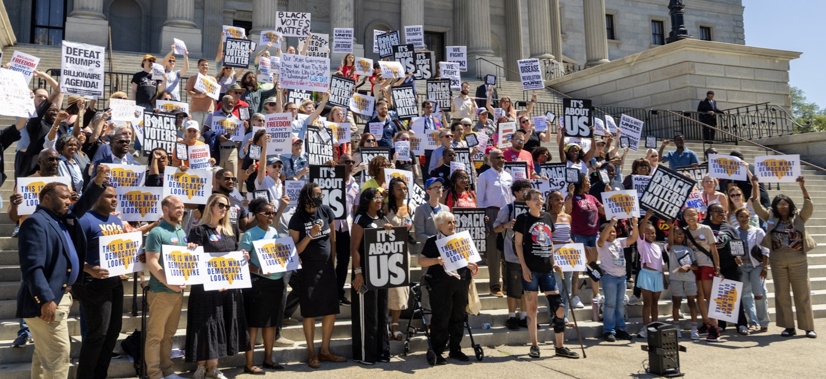A diverse crowd of protesters on the Statehouse steps holding signs protesting against redistricting