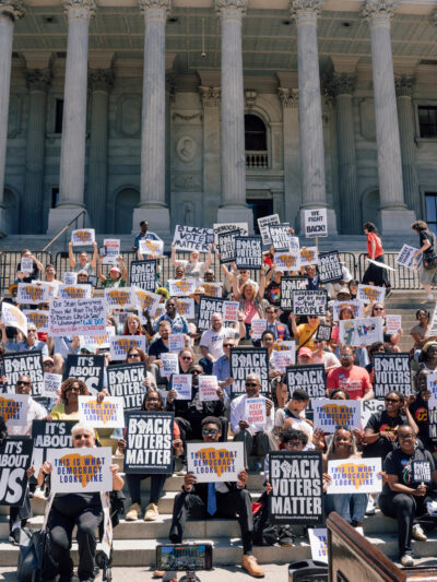 A crowd of protesters on the Statehouse steps. Signs say "This is what democracy looks like" and "Black voters matter."