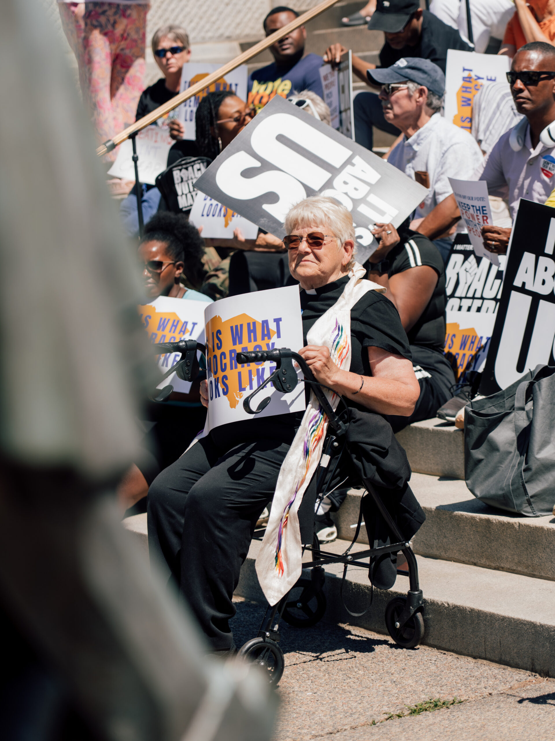 A woman seated at a rally holding a sign that says This is What Democracy Looks Like