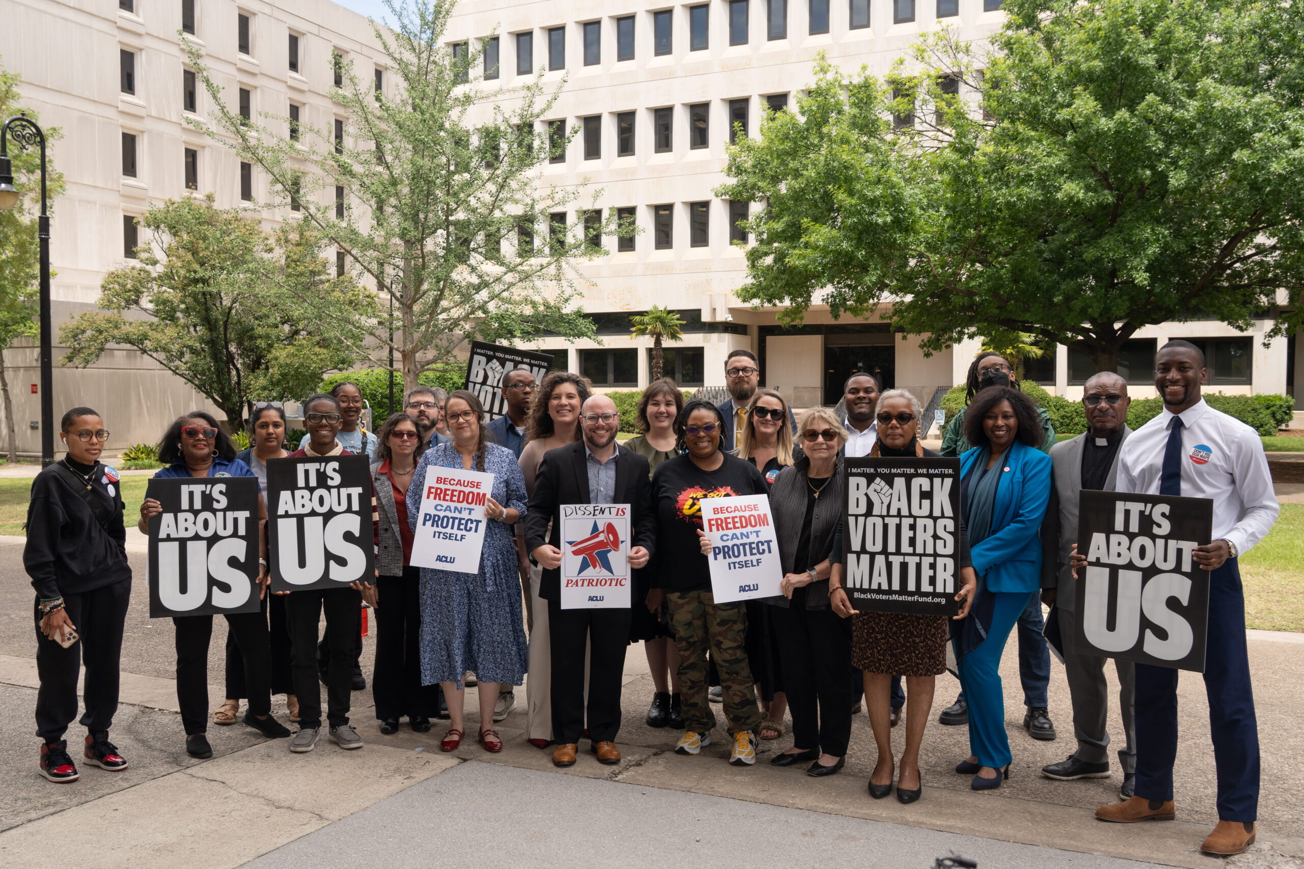 A press conference on the Statehouse grounds. Participants are holding signs that say "Black Voters Matter," "It's About US," and "Dissent is Patriotic"