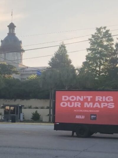 A billboard truck drives past the South Carolina Statehouse. A sign on the truck says "Don't rig our maps. aclusc.org/rigged"