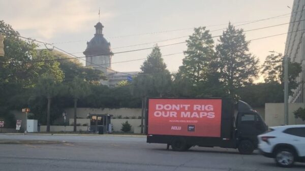 A billboard truck drives past the South Carolina Statehouse. A sign on the truck says "Don't rig our maps. aclusc.org/rigged"