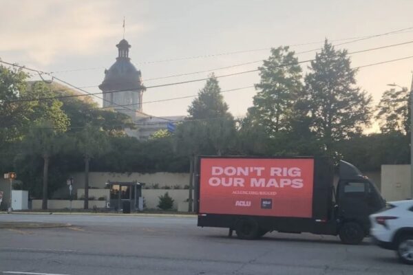 A billboard truck drives past the South Carolina Statehouse. A sign on the truck says "Don't rig our maps. aclusc.org/rigged"