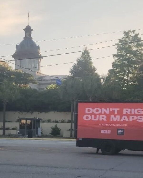 A billboard truck drives past the South Carolina Statehouse. A sign on the truck says "Don't rig our maps. aclusc.org/rigged"