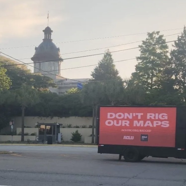 A billboard truck drives past the South Carolina Statehouse. A sign on the truck says "Don't rig our maps. aclusc.org/rigged"