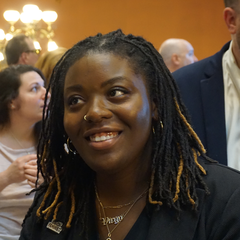 A headshot of Courtney in the State House lobby. She has her hair in braids and is wearing a dark suit jacket and a gold necklace that says "Virgo"
