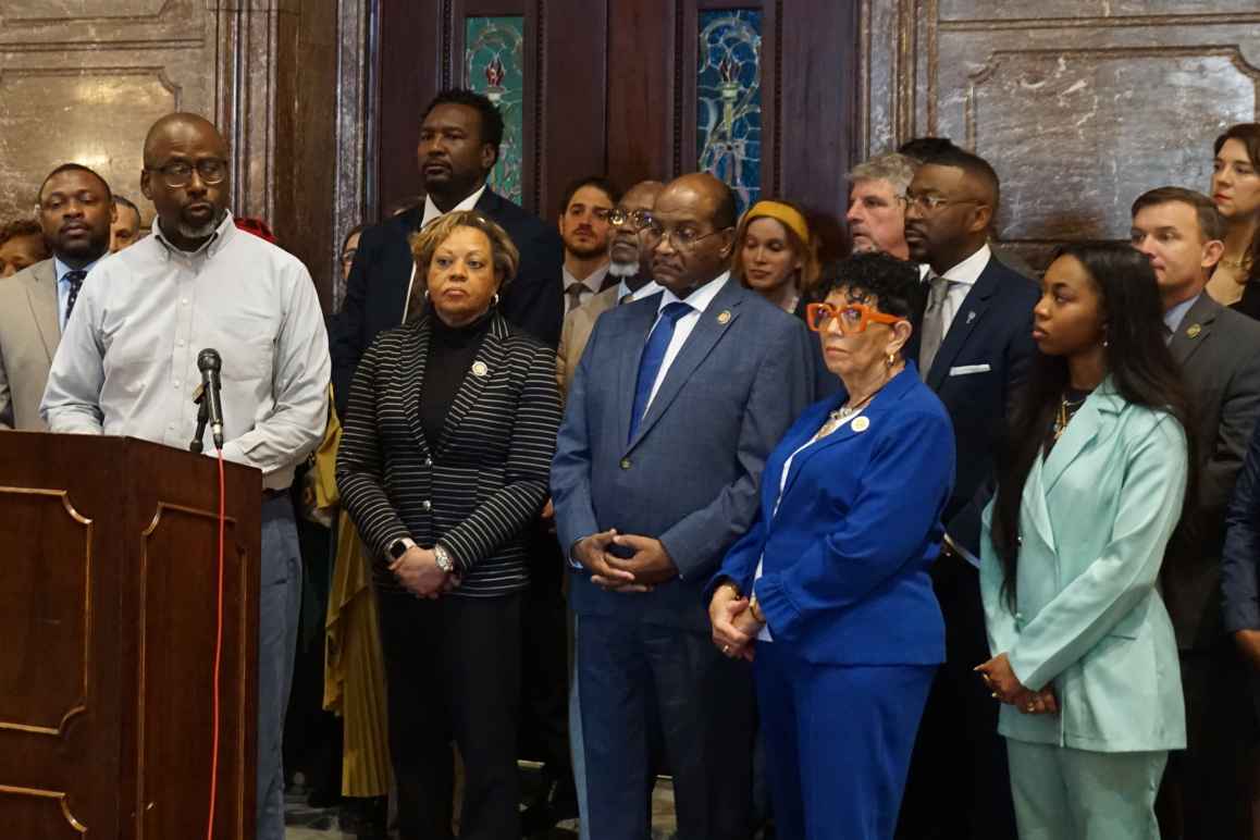 A crowd of people in businesswear stands behind a speaker at a lectern in the South Carolina State House lobby