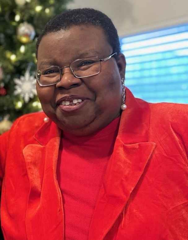 Tish Gotell Faulks in a bright red suit jacket and red shirt. She has a close cropped haircut, wire-rimmed glasses, and pearl earrings.