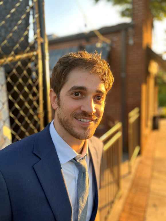 A photo of Josh Malkin in a  navy blue suit standing in the sunlight beside a chain link fence