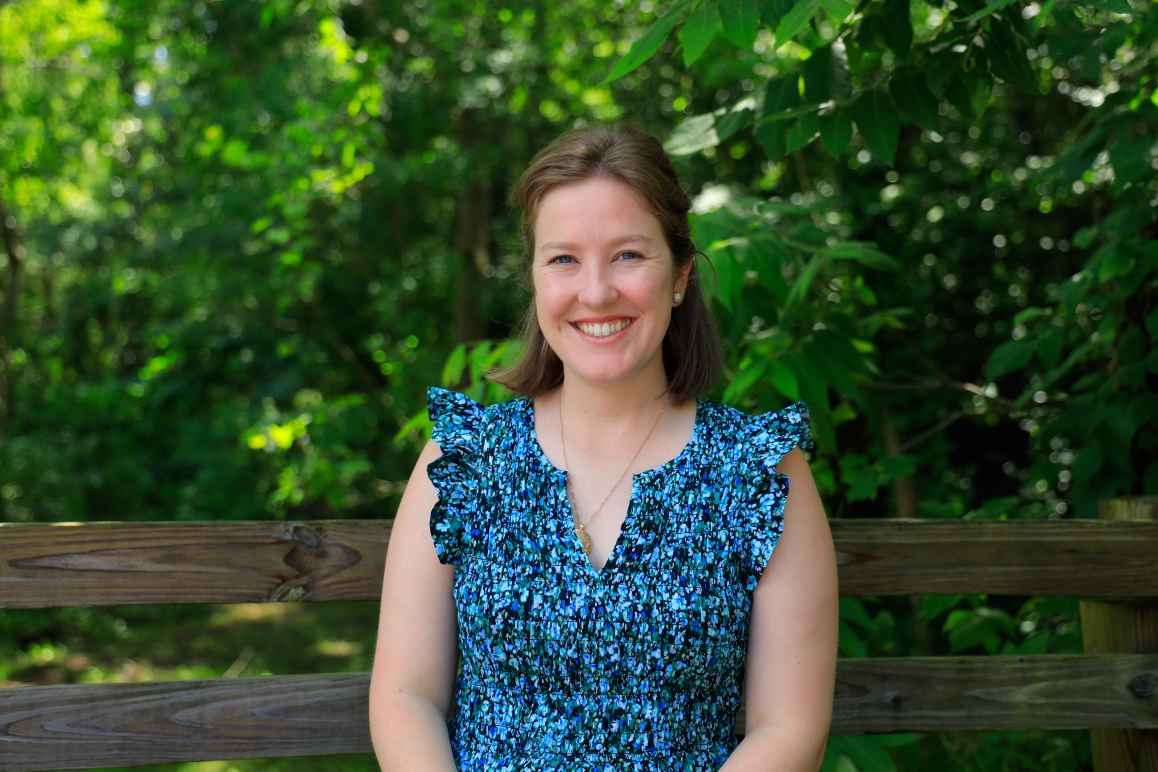 Meredith is smiling and wearing a blue blouse in front of a lush green background.