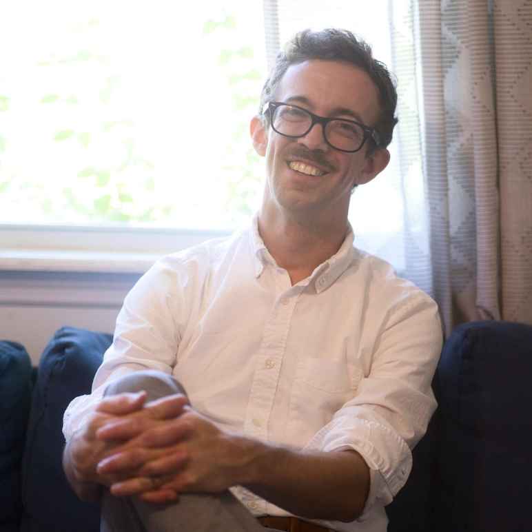 Paul Bowers in a white dress shirt on a blue couch. He is smiling and his hands are folded on his knee.