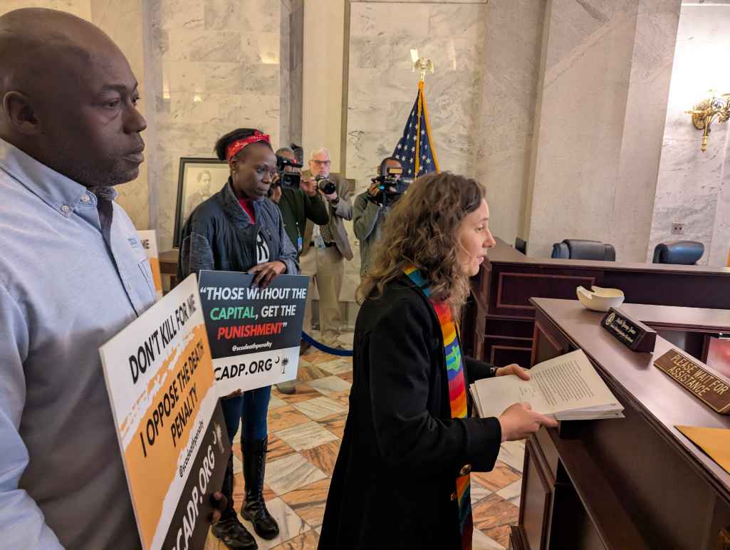 Petitioners, including a white woman in minister's garments and a Black man in a dress shirt, deliver petitions at a desk in the State House lobby.