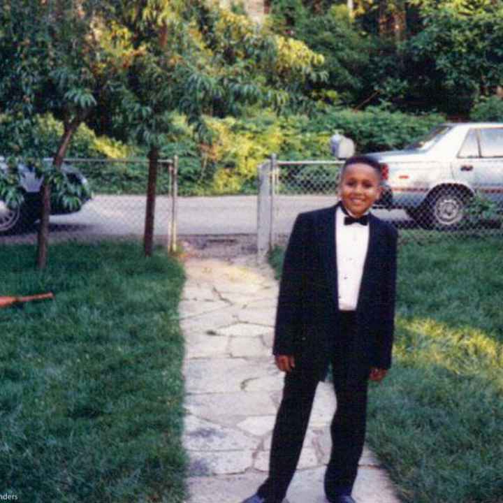 A young Black boy stands in a front yard smiling and wearing a tuxedo