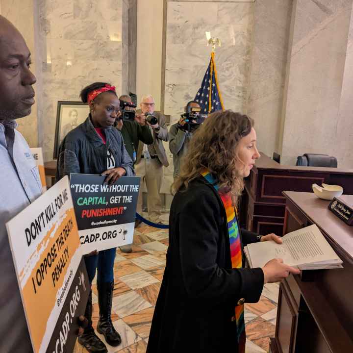 Petitioners, including a white woman in minister's garments and a Black man in a dress shirt, deliver petitions at a desk in the State House lobby.