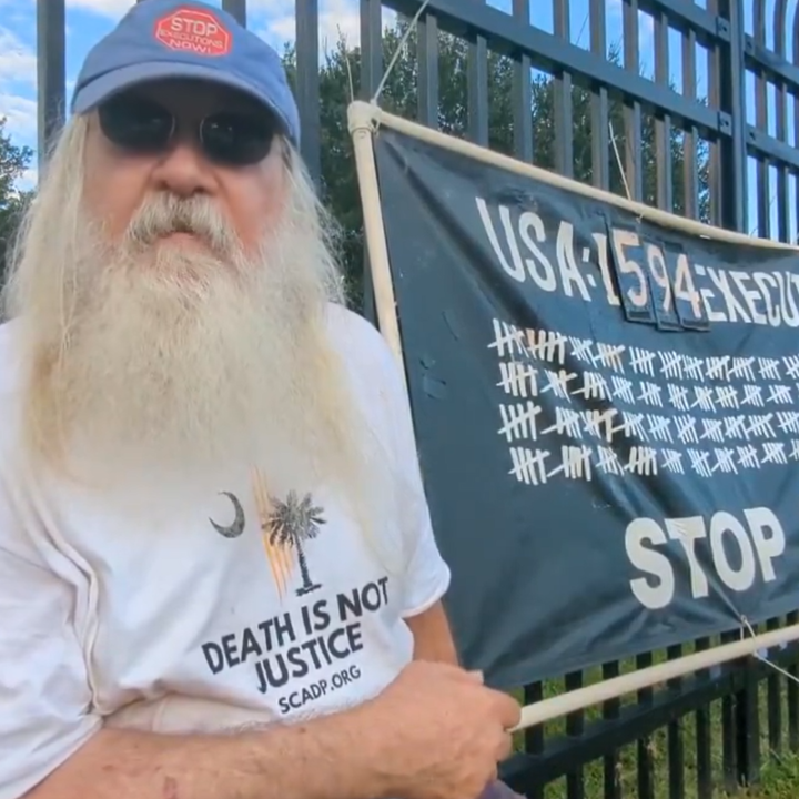 A man with a long white beard sits beside a black banner that reads, "USA: 1594 Executions 1977 - Present. Stop Executions!"