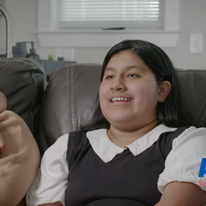 Skyler, a 13-year-old boy, smiles while talking. He is seated on a brown leather couch beside his mother.